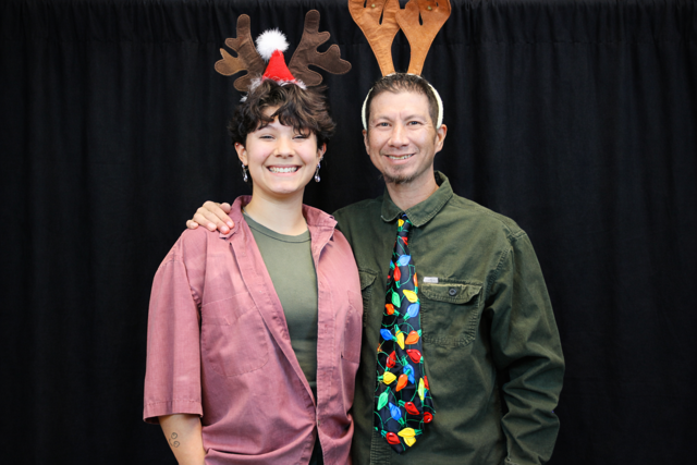 Two guests smiling in front of a black photo booth backdrop wearing festive holiday reindeer antlers.