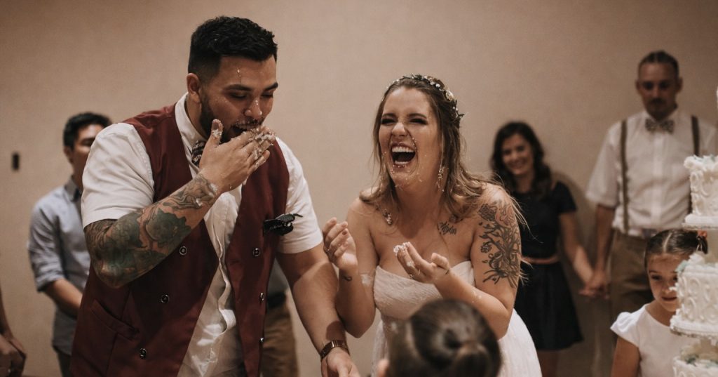 Joyful bride and groom laughing during a fun cake smash at their Utah wedding reception, covered in cake and celebrating with guests.
