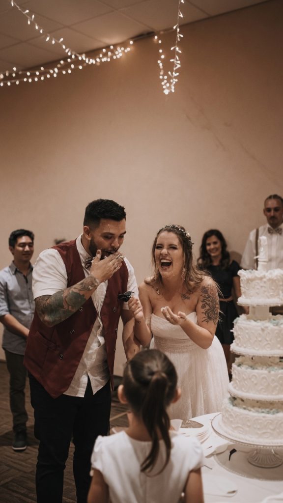 Joyful bride and groom laughing during a fun cake smash at their Utah wedding reception, covered in cake and celebrating with guests.